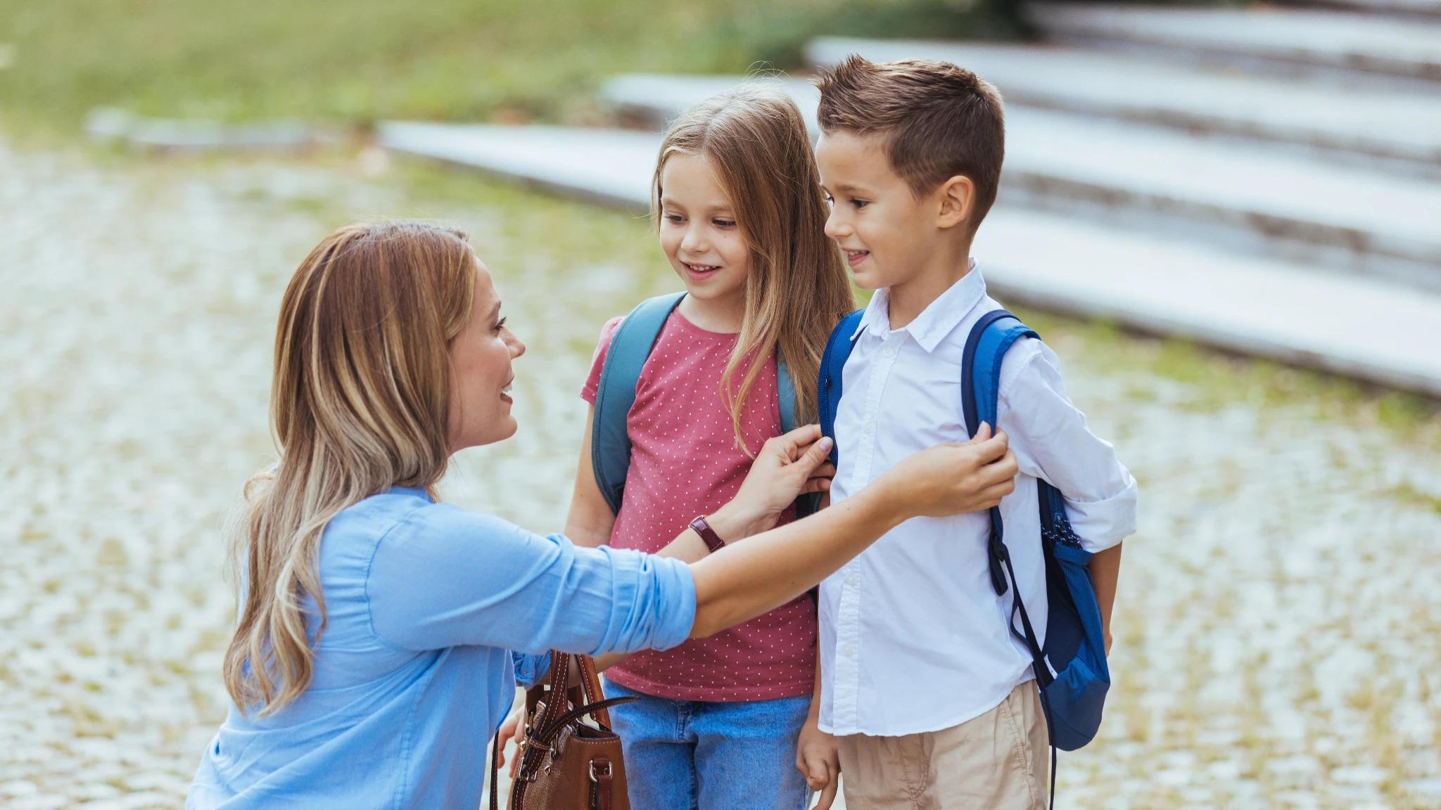 An adult woman speaking with two young children wearing backpacks outdoors, appearing to check on them before school. The children look attentive and reassured, suggesting a supportive and pr