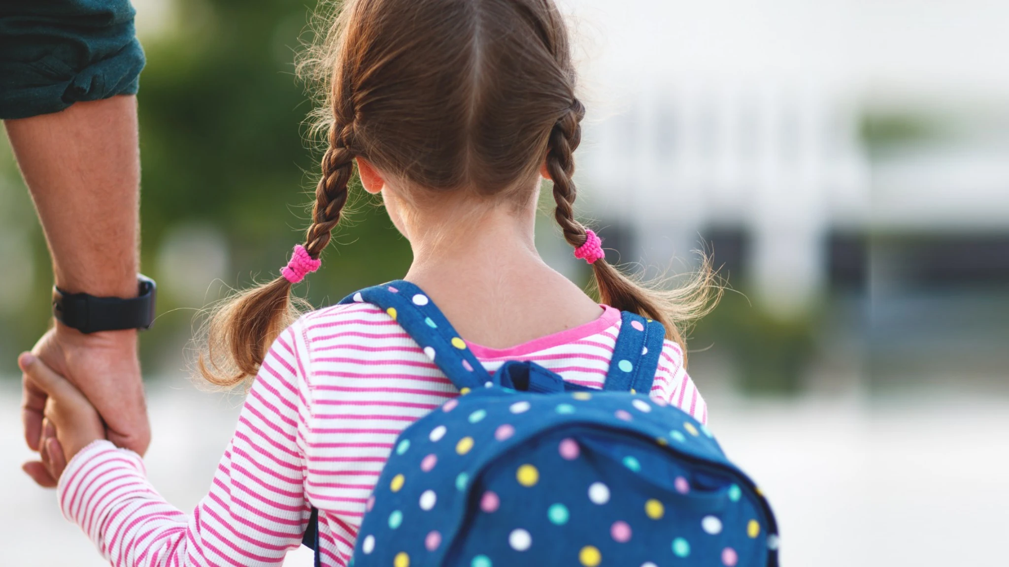 A girl with a polka-dot backpack seen from behind, walking beside an adult